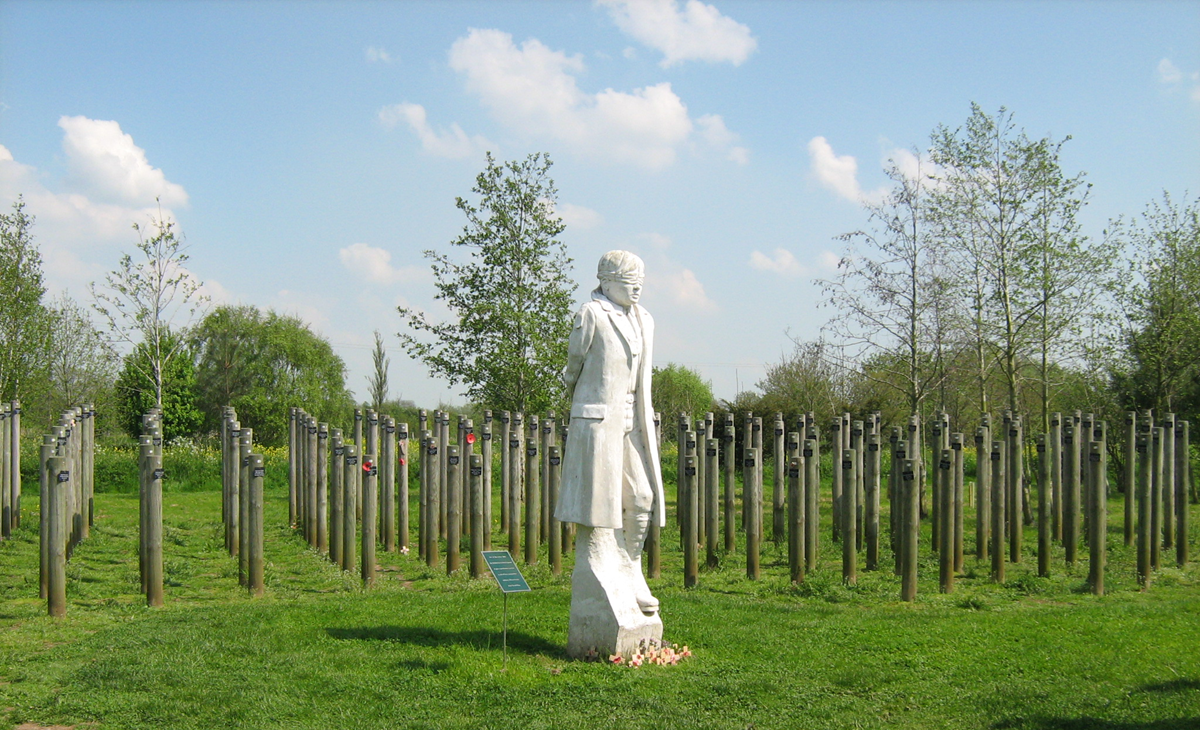 A white statue of a person in a coat stands in a grassy field surrounded by young trees and wooden posts, under a blue sky with scattered clouds.