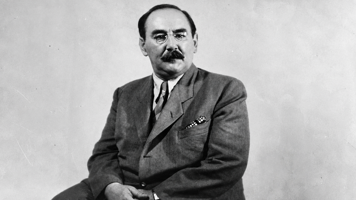 A man with a mustache and glasses, dressed in a suit and tie, sits against a plain, light-colored background, looking slightly to the side with a neutral expression.