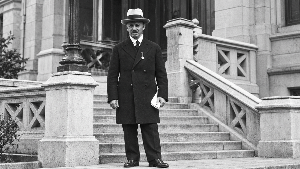 A man in a dark coat and hat stands in front of stone steps leading to a large building. He holds a paper in one hand and wears a medal around his neck. The scene appears to be from the early 20th century.