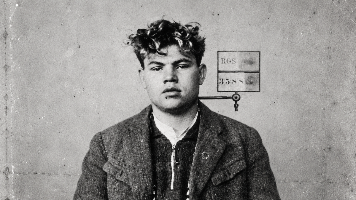 Black and white photo of a young man with curly hair wearing a coat and shirt, sitting against a plain wall with a small sign featuring letters and numbers behind him.