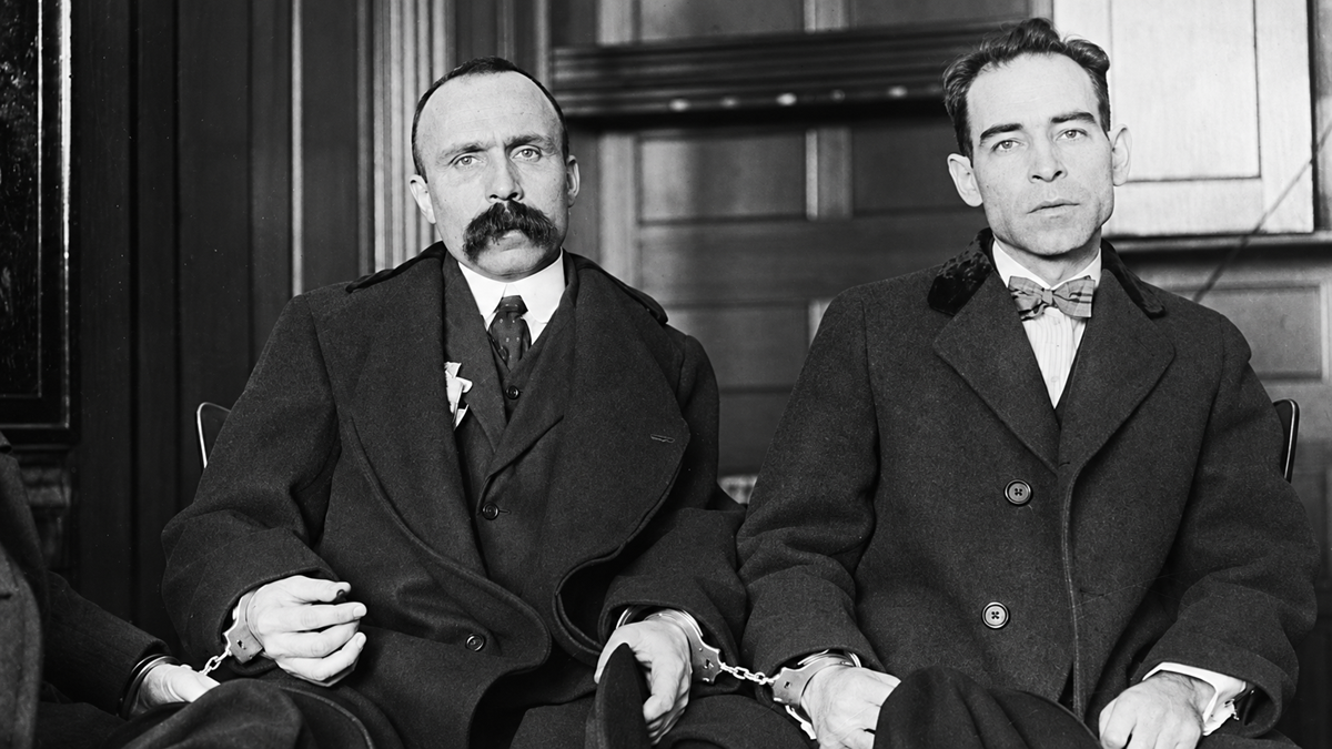 Two men in suits sit side by side, handcuffed together, in what appears to be a courtroom. One man has a large mustache, and the other wears a bow tie. Both look serious and somber.