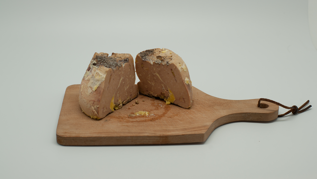Two pieces of foie gras sit on a small wooden cutting board with a handle and a leather loop, against a plain light background.