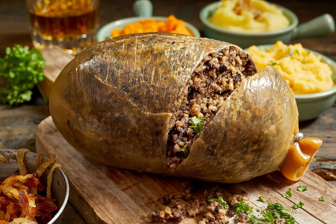 A traditional Scottish haggis, cut open to reveal its filling of oats, meat, and spices, sits on a wooden board with mashed potatoes and vegetables in the background.
