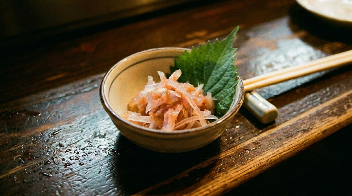 A small ceramic bowl with thinly sliced sashimi and a green shiso leaf, placed on a rustic wooden table next to a pair of chopsticks.