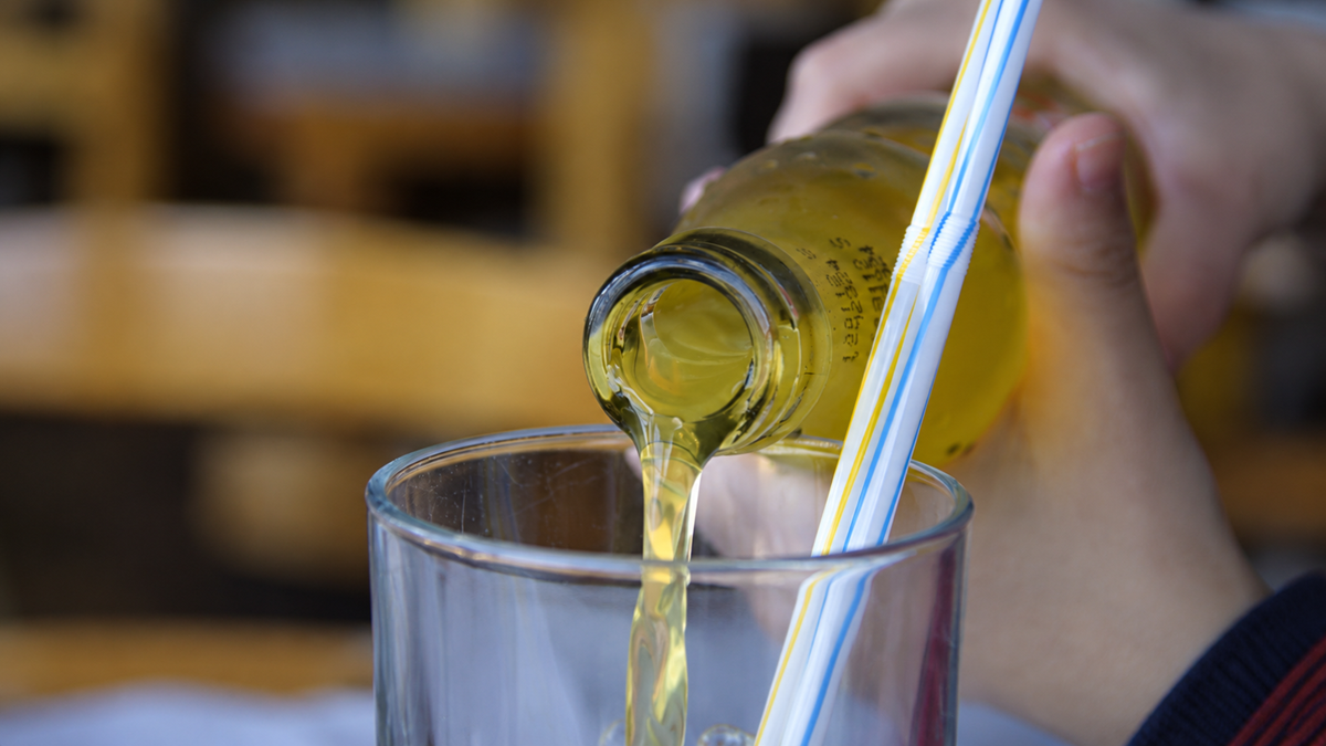 A close-up of a hand pouring yellow soda from a glass bottle into a clear glass with two striped straws. The background is blurred and features wooden chairs.