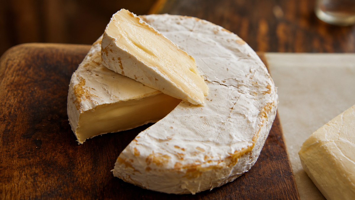 A wheel of brie cheese with a wedge cut out, showing its creamy interior. The cheese sits on a wooden board with a rustic background.