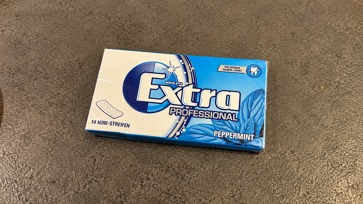 A pack of Wrigley’s Extra Professional Peppermint gum with 14 mini strips, placed on a brown textured surface. The packaging is blue and white with mint leaves pictured.