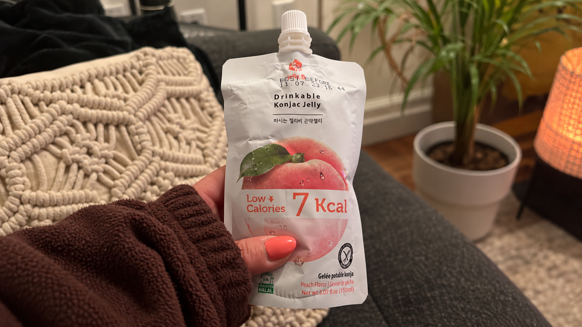 A hand holds a pouch of drinkable konjac jelly with a peach image on the front and “7 Kcal” label. Background shows a potted plant, lamp, and part of a sofa.