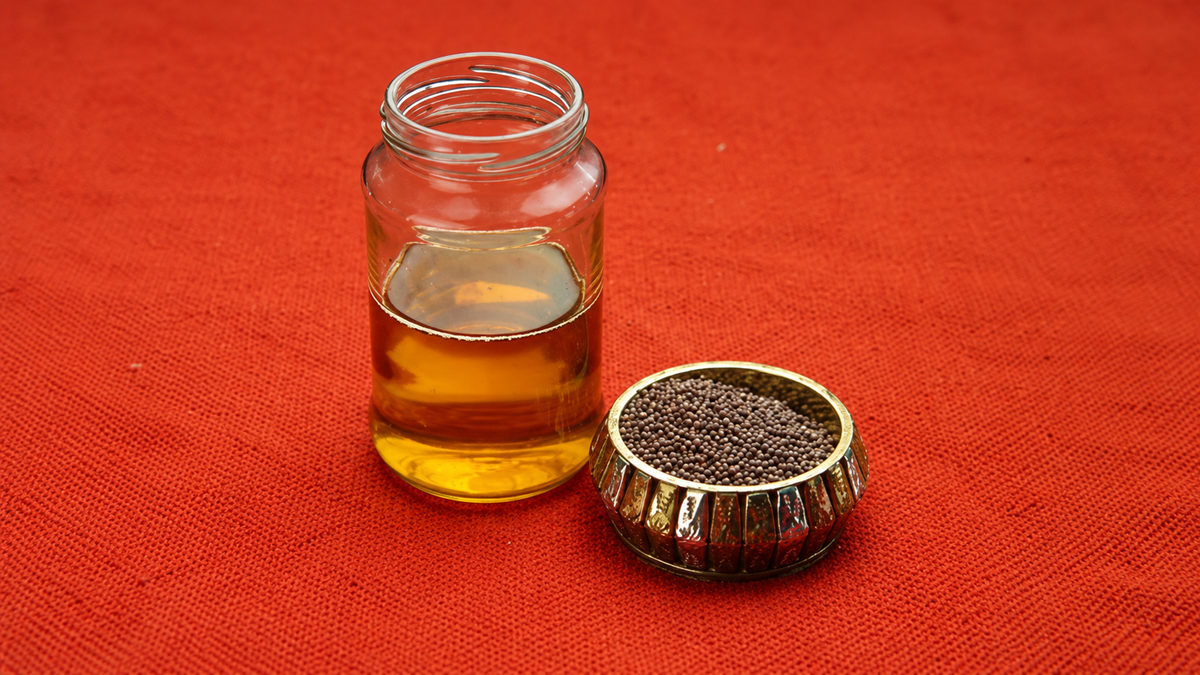 A glass jar of yellow oil and a small metal bowl filled with black mustard seeds sit on a textured red surface.