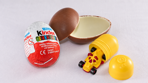 A Kinder Surprise egg is shown, with its chocolate shell broken open to reveal a yellow capsule containing a small toy race car.