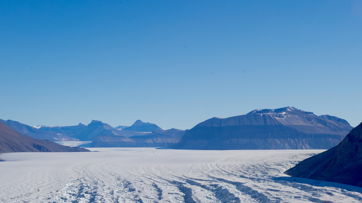 Snow-covered mountains and a glacier under a clear blue sky, with deep crevasses visible in the foreground and distant rugged peaks in the background.