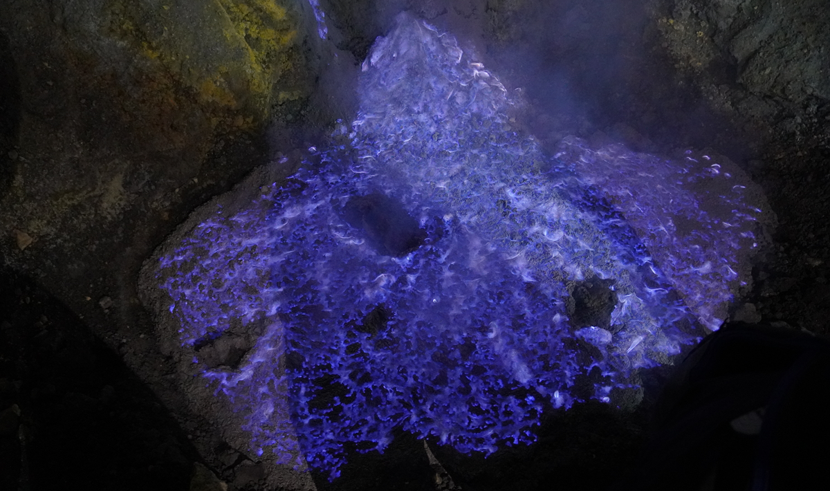 Blue flames rise from rocky ground at night, likely caused by burning sulfur gas. The vibrant blue and purple hues stand out against the dark surrounding area.