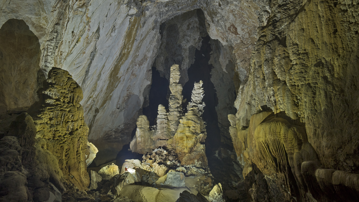 A vast, dimly lit cave with tall stalagmites and rugged stone formations. A small person with a flashlight stands near the center, emphasizing the massive scale of the cavern.