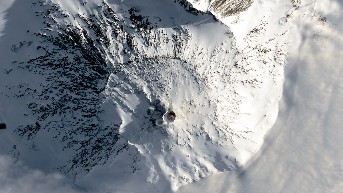A snow-covered volcano seen from above, with a dark crater at the center emitting a small amount of smoke and a faint red glow. Rugged, icy slopes surround the crater under bright daylight.
