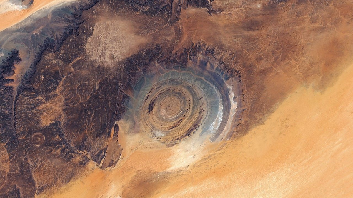 Aerial view of the Richat Structure, a circular geological formation in the Sahara Desert, featuring concentric rings in shades of brown, tan, and gray surrounded by desert sands.