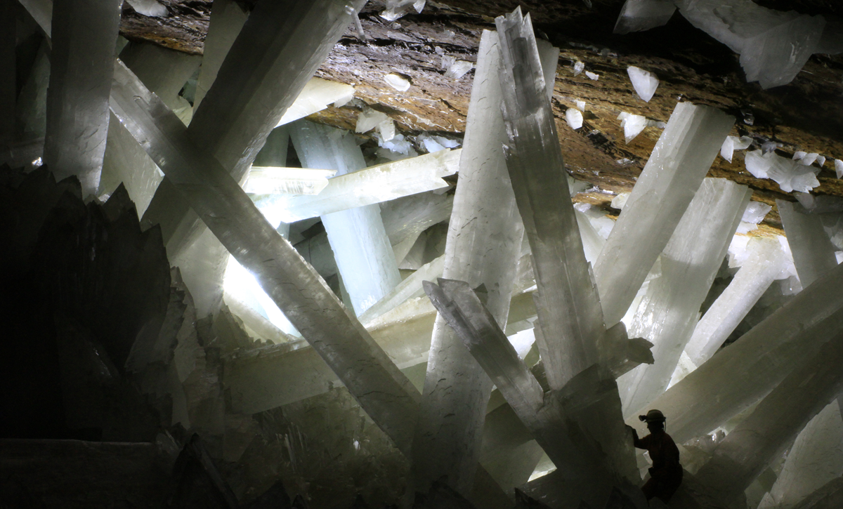 A person wearing a helmet stands among enormous, clear gypsum crystals inside a dimly lit cave, with light shining through the large crystal formations.