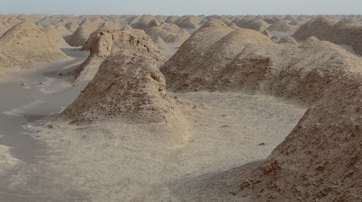 A desert landscape features numerous sandy, rounded mounds under a hazy sky. The terrain appears dry, with ridges and shadows emphasizing the texture of the earth. No vegetation or signs of life are visible.