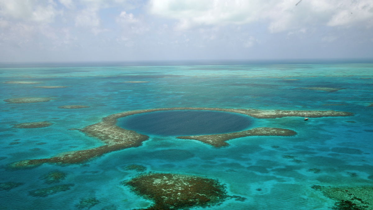 Aerial view of the Great Blue Hole, a large, circular marine sinkhole surrounded by turquoise water and coral reefs in the ocean. A small boat is visible near the edge.