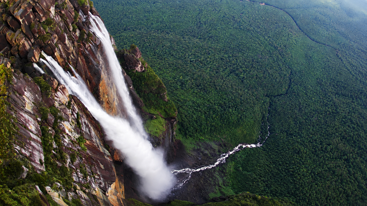 A tall waterfall cascades from a rocky cliff into a lush green forest below, with a winding river visible through the dense trees.