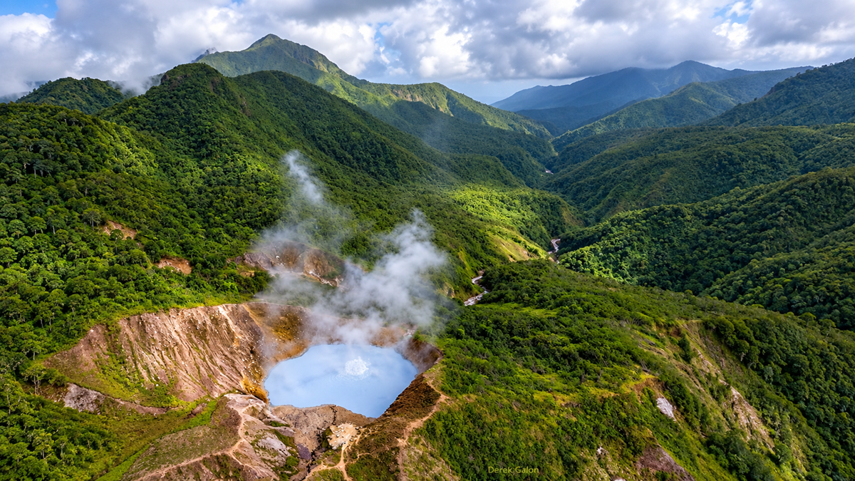 Aerial view of a steaming volcanic crater lake surrounded by lush green mountains and dense forest under a partly cloudy sky.