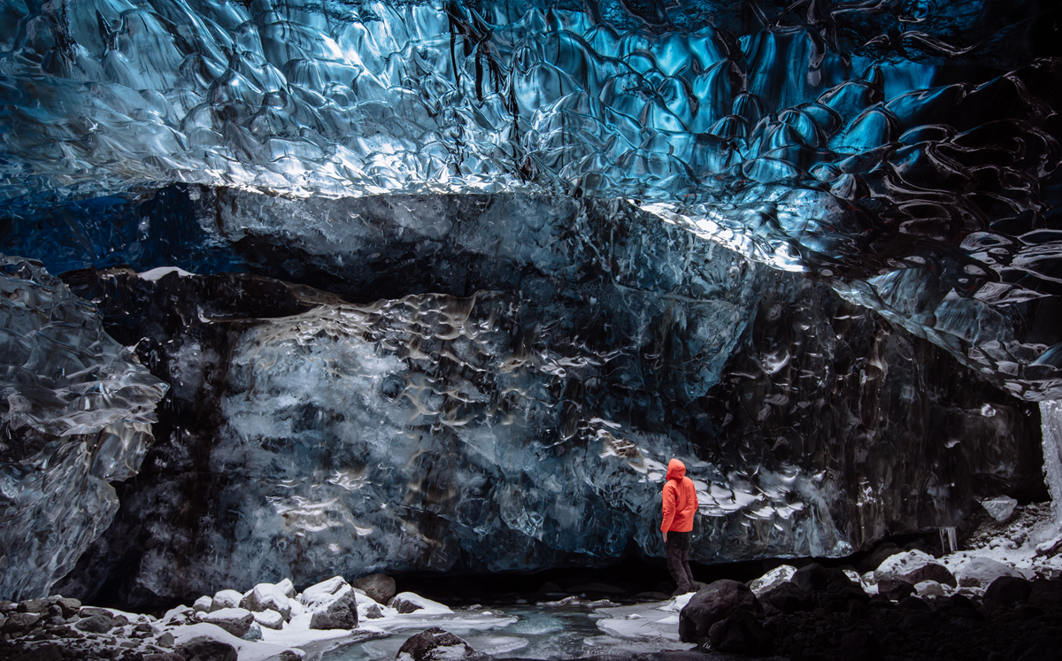 A person in an orange jacket stands inside an icy blue cave, surrounded by textured, glistening ice walls and a partially frozen ground.