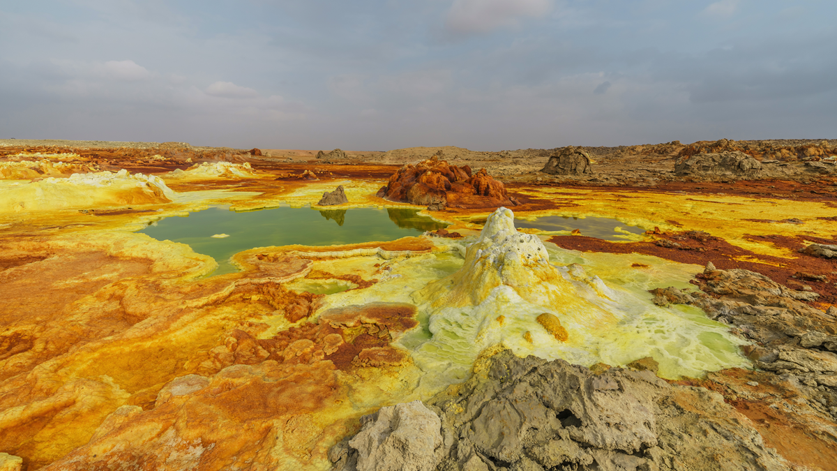 Extreme places. A colorful geothermal landscape with yellow, green, and orange mineral deposits, small pools of greenish water, and rugged terrain under a cloudy sky.