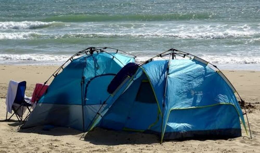Two blue tents and a chair set up on a sandy beach, with waves and the ocean visible in the background under a bright, clear sky.