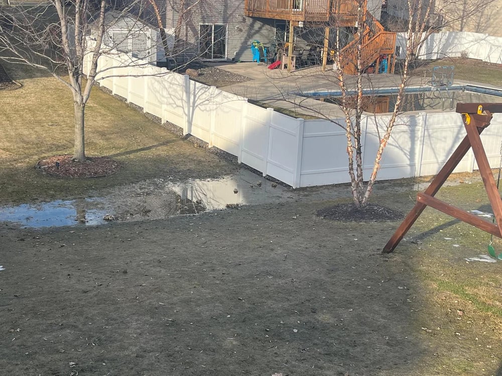 A backyard with patchy grass, leafless trees, a swing set, a white fence, a large puddle near the fence, and a wooden deck with outdoor toys.