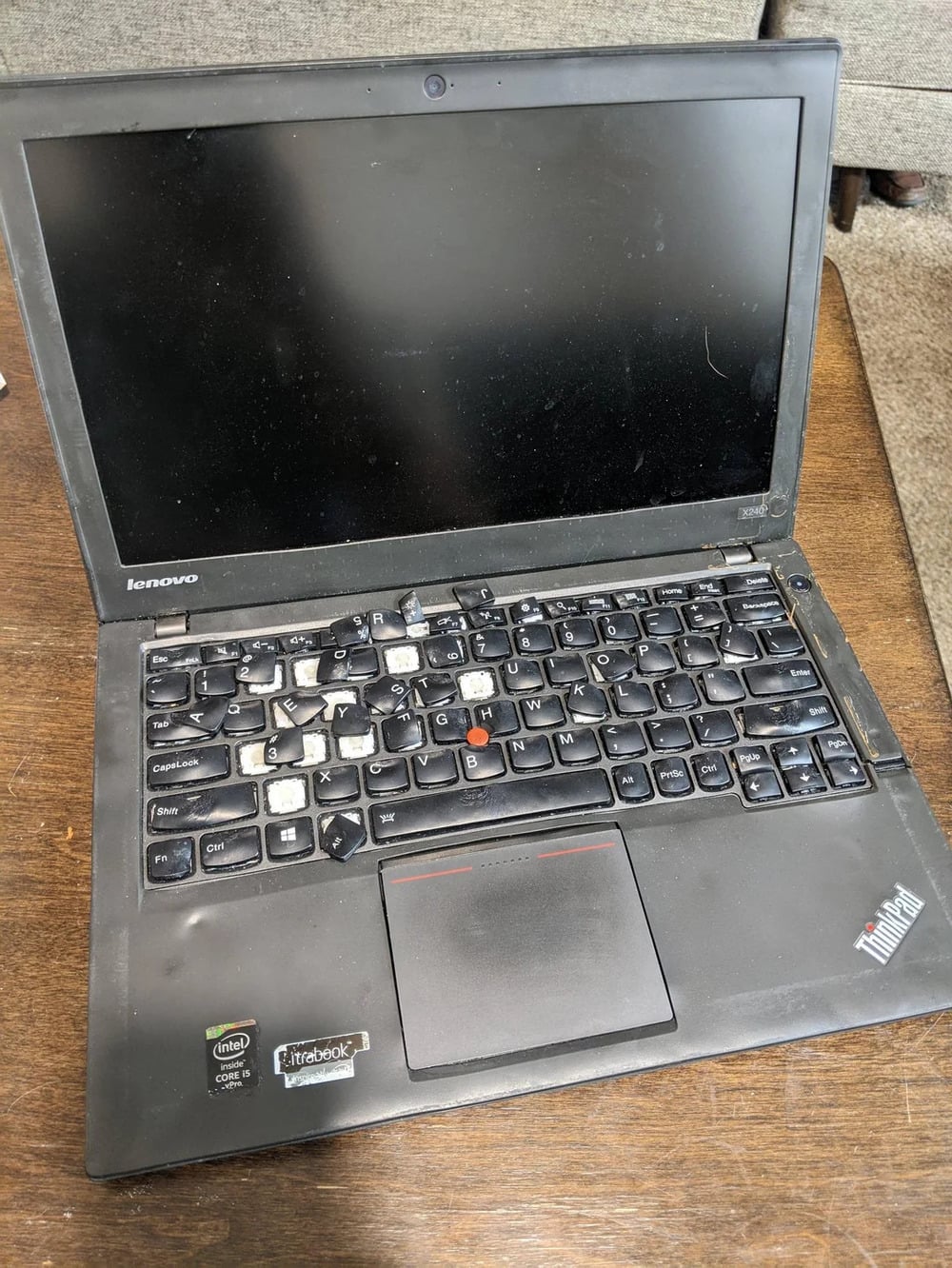 A black Lenovo ThinkPad laptop with several worn and missing keys sits on a wooden surface. The screen is off, and the laptop appears used and slightly damaged.