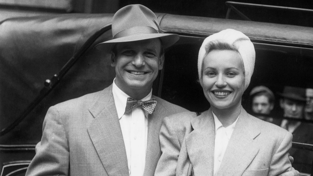 A smiling man in a suit, bow tie, and hat stands beside a smiling woman in a suit and turban-style headscarf. They pose together in front of a classic car, with people visible in the background.