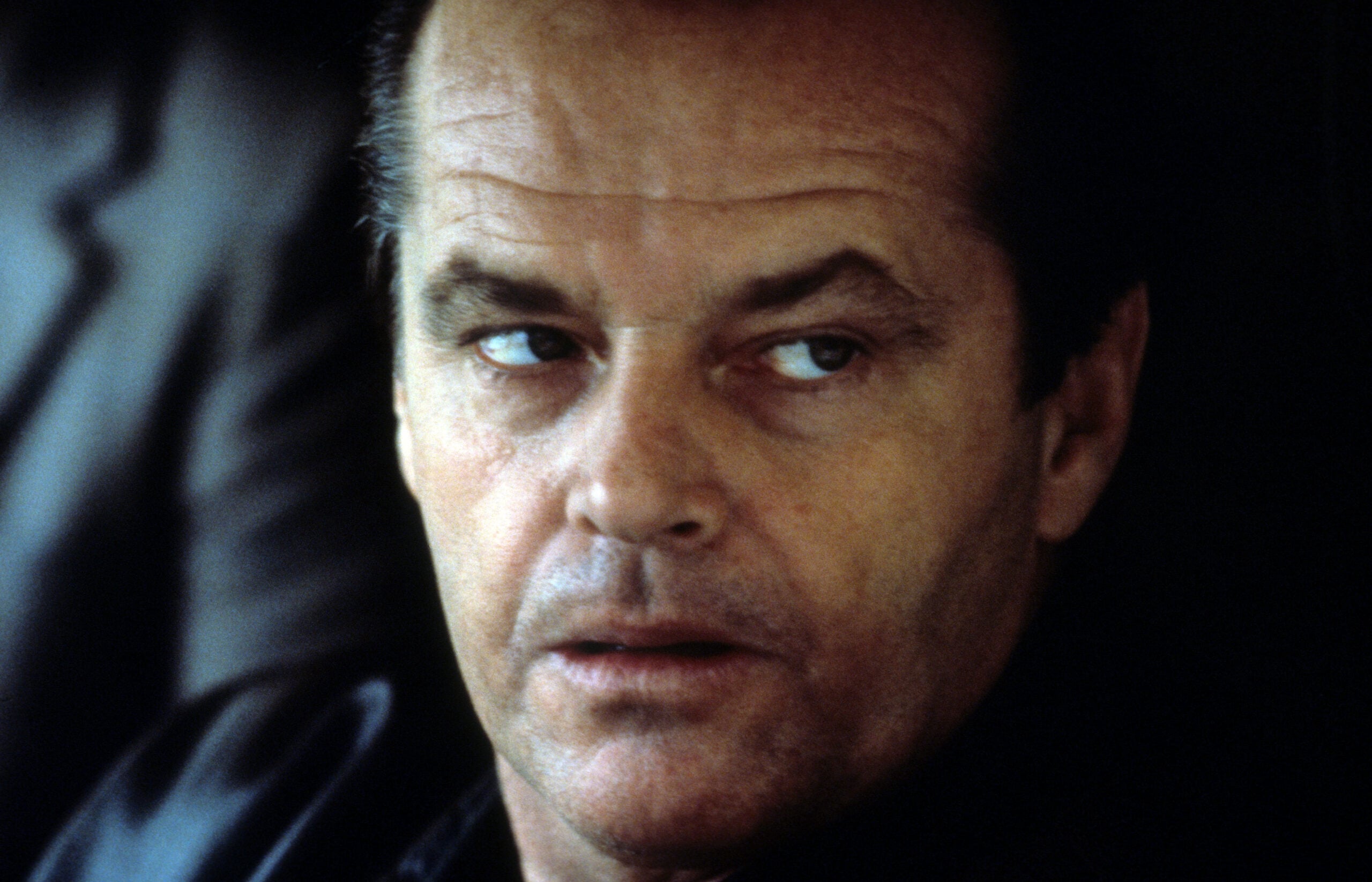 A man with slicked-back hair and a serious expression looks slightly to the side. He wears a dark jacket and is lit from the front, with part of another person’s shoulder visible in the background.