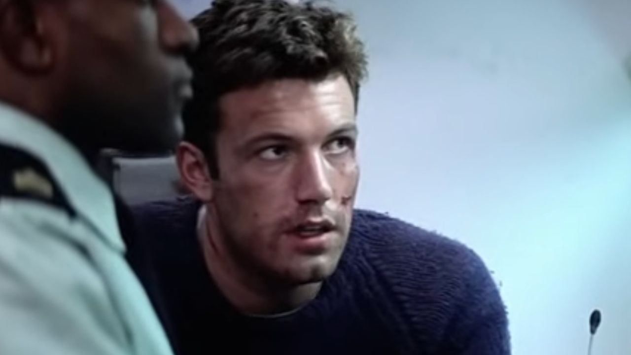 A man with short brown hair and a bruised face, wearing a dark sweater, sits next to a person in a uniform in a dimly lit room, looking concerned.