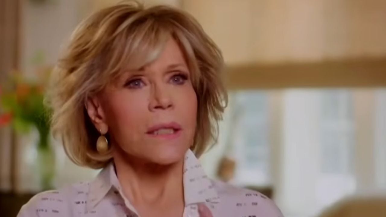 A woman with short blonde hair and gold earrings is indoors, looking slightly to the side. She wears a light-colored collared shirt. The background is softly blurred.