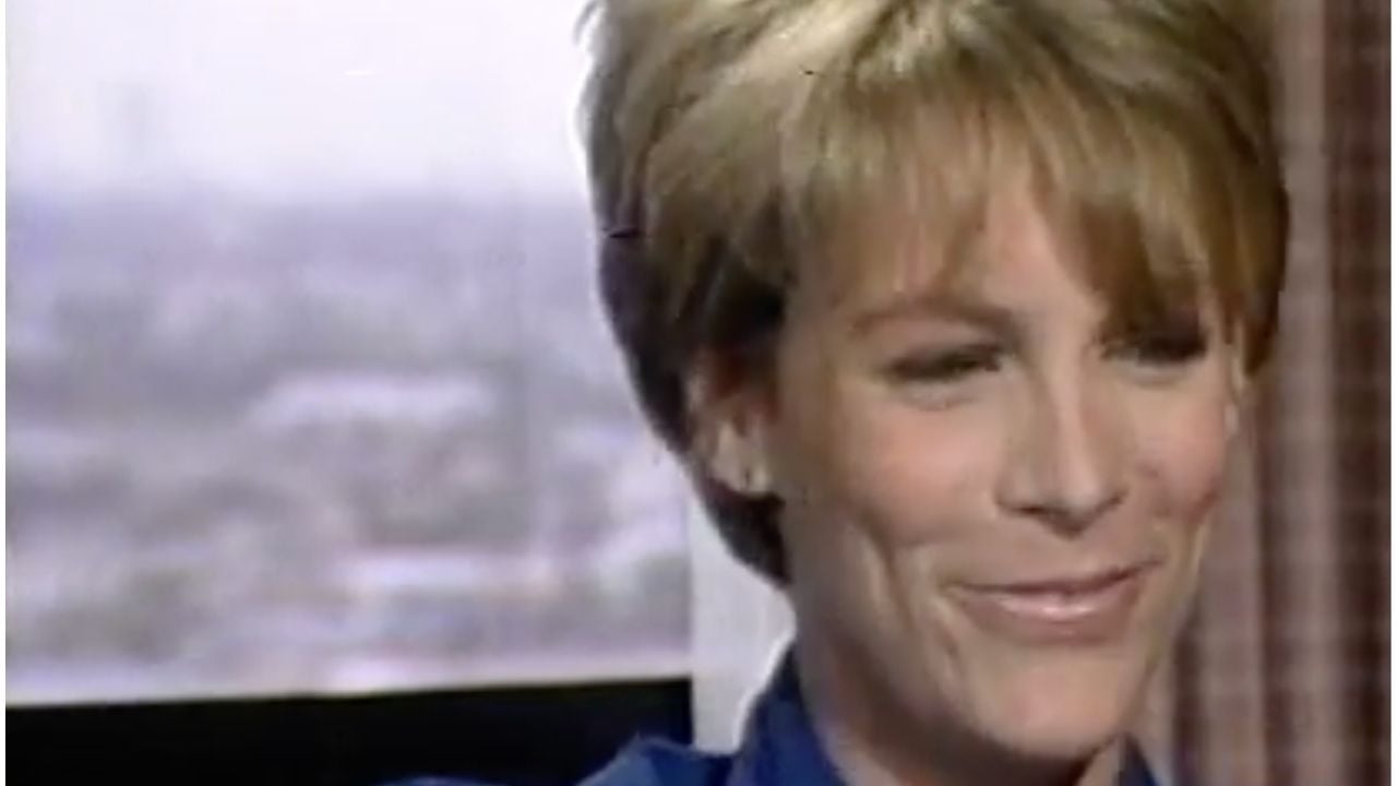 A person with short, light brown hair is smiling softly, wearing a blue collared shirt. The background is blurred, showing a window with an outdoor city view and a curtain on the right.