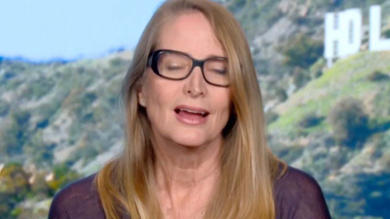 A woman with long blonde hair and glasses is speaking with her eyes closed. The Hollywood sign and a green hillside are visible in the background.