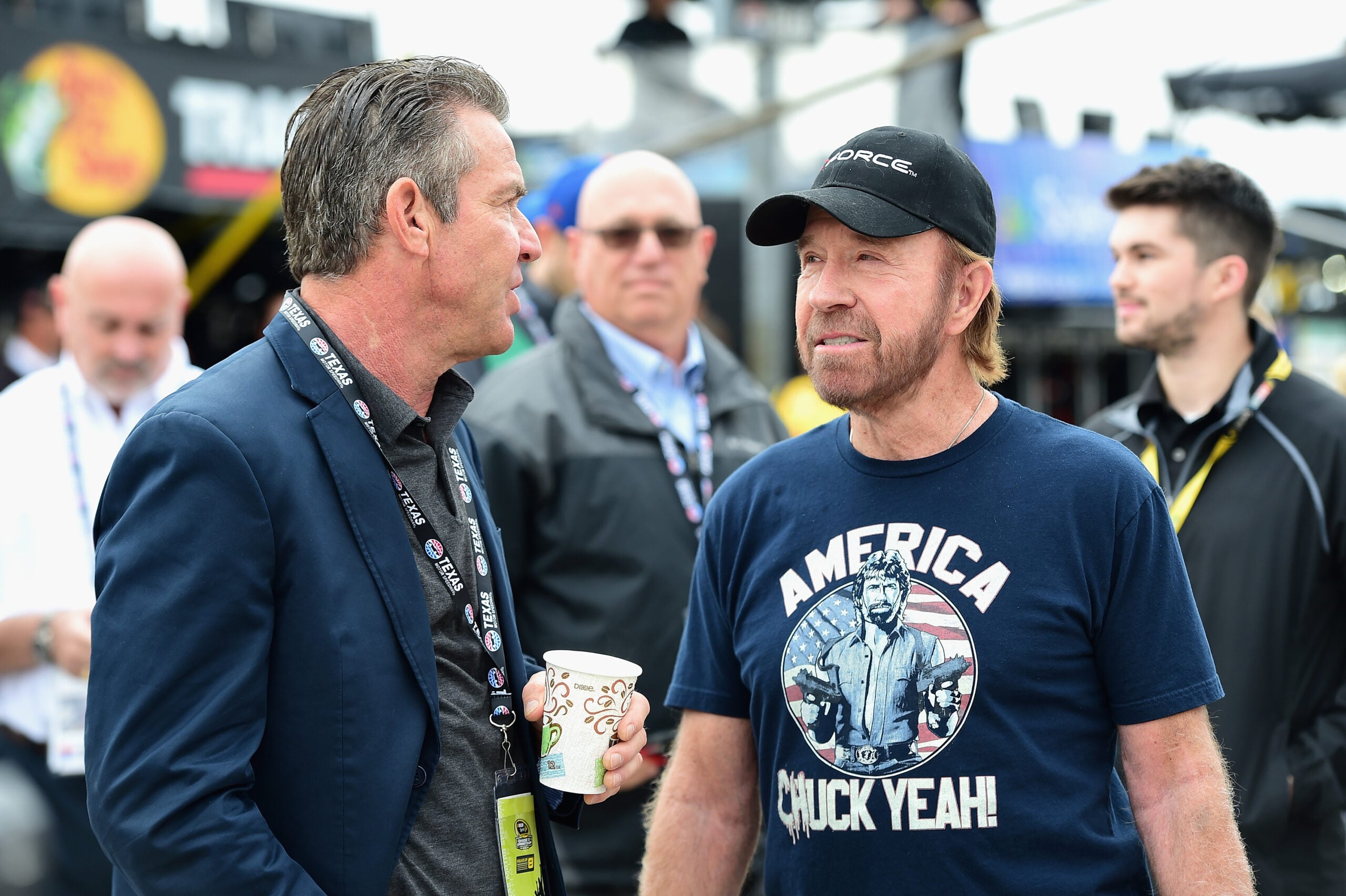 Two men talk at an outdoor event. One wears a navy blazer and holds a coffee cup. The other wears a cap and a shirt that says “America Chuck Yeah!” More people are blurred in the background.