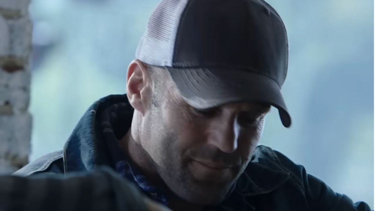 A man wearing a dark baseball cap and jacket looks down with a slight, thoughtful expression. The background is softly blurred with cool tones.