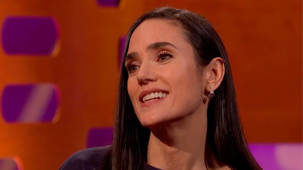 A woman with long dark hair smiles while seated against an orange background with purple accents. She is wearing a dark top and small earrings.