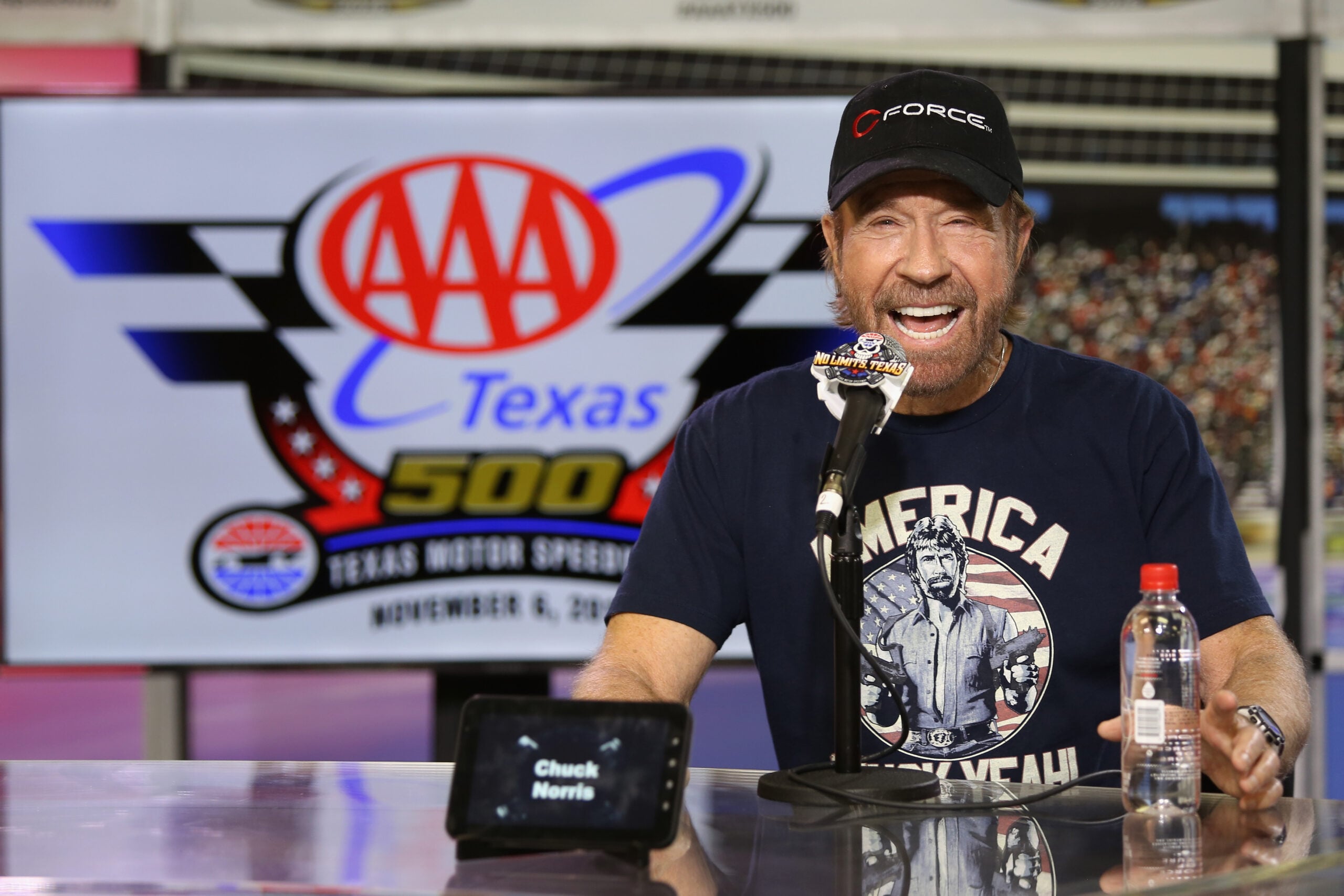 A man with a beard and cap speaks into a microphone at a press event for the AAA Texas 500, with event signage in the background and a water bottle and nameplate on the table.