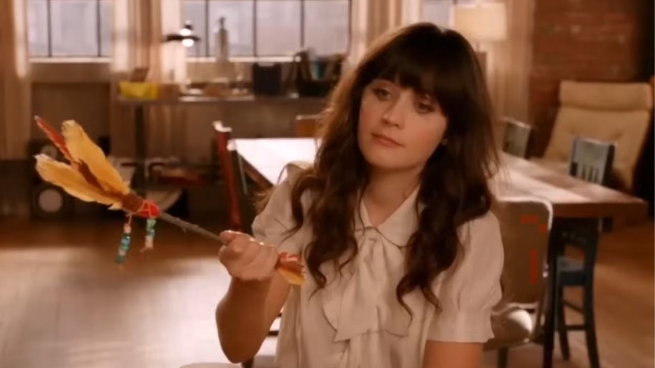 A woman with long dark hair sits indoors holding a feather duster, wearing a light-colored blouse, looking off to the side with a neutral expression. The room is warmly lit, with a table and chairs in the background.