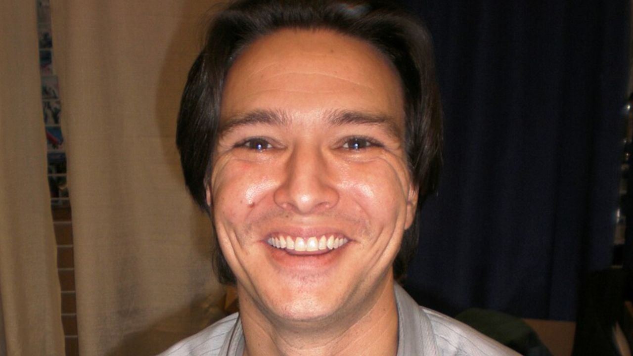 A person with straight brown hair and a wide smile stands indoors in front of beige and dark blue curtains.