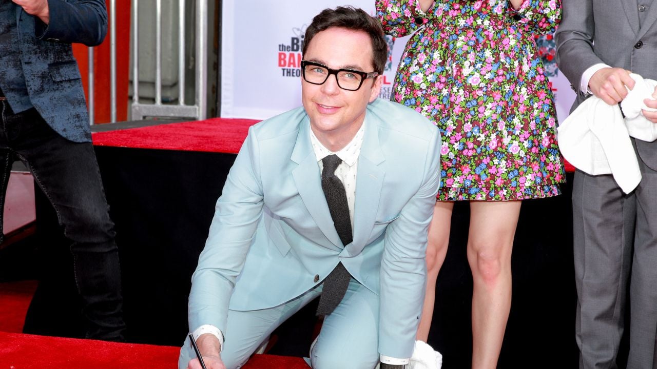 A man in a light blue suit and black glasses kneels on a red carpet, smiling while signing an autograph. A woman in a colorful floral dress stands behind him.