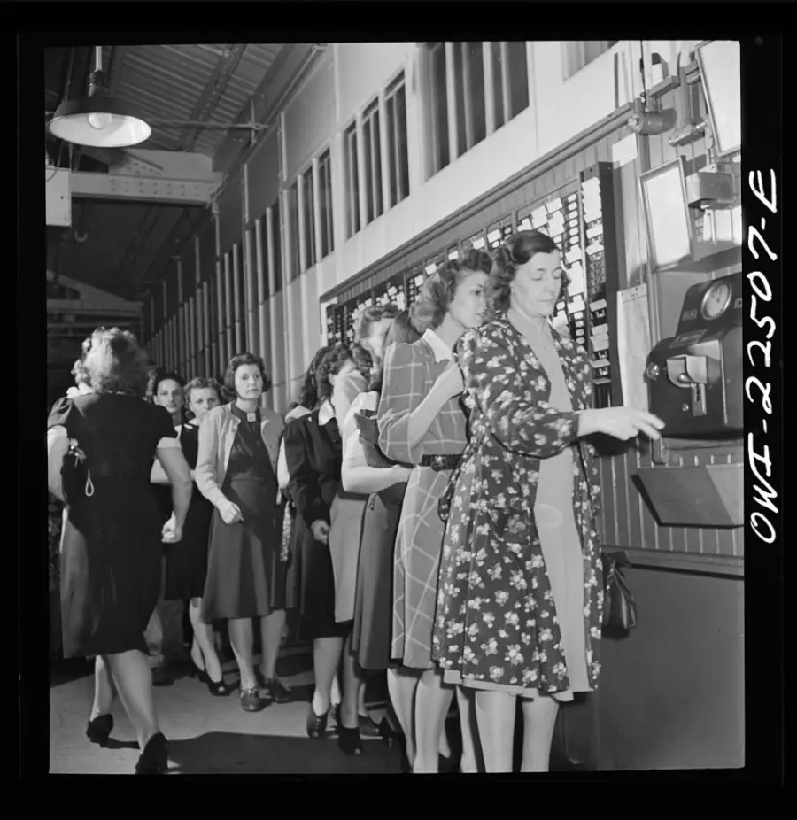 Women in 1940s attire stand in line to clock in or out at a timecard machine in a factory hallway, with an industrial setting visible in the background.