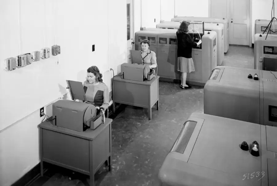 Three women work at vintage data entry machines in a large room, using punch card equipment and tabulators, likely from the mid-20th century. The setting appears to be an office or data processing center.