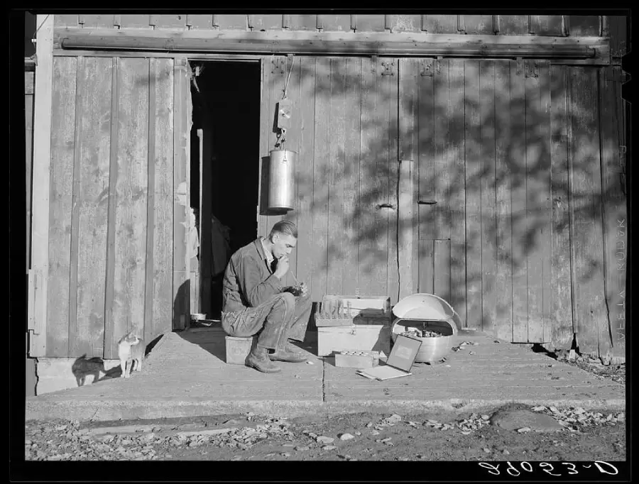 A man sits on a box in front of a wooden building, examining something in his hands. Various tools and containers are nearby. A small dog stands at the edge of the shadow by the open door.