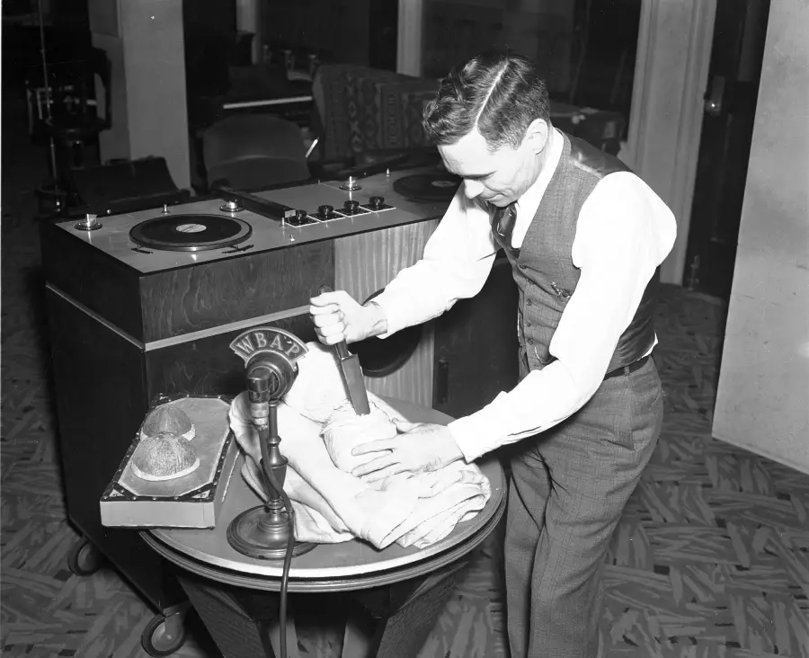 A man in a vest and tie uses a knife on a cabbage in front of a WBAI microphone, likely creating sound effects, with audio equipment and a record player in the background.