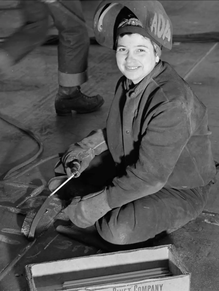 A woman in protective gear and a welding helmet sits on the floor, smiling while holding welding equipment. A box and another person’s legs are visible nearby.