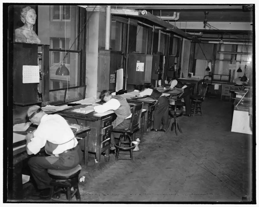 Four men sit at drafting tables working on papers in a large, industrial-style room with high windows and hanging lamps. A calendar and a bust are visible on the wall to the left. The space appears to be a mid-20th-century office.