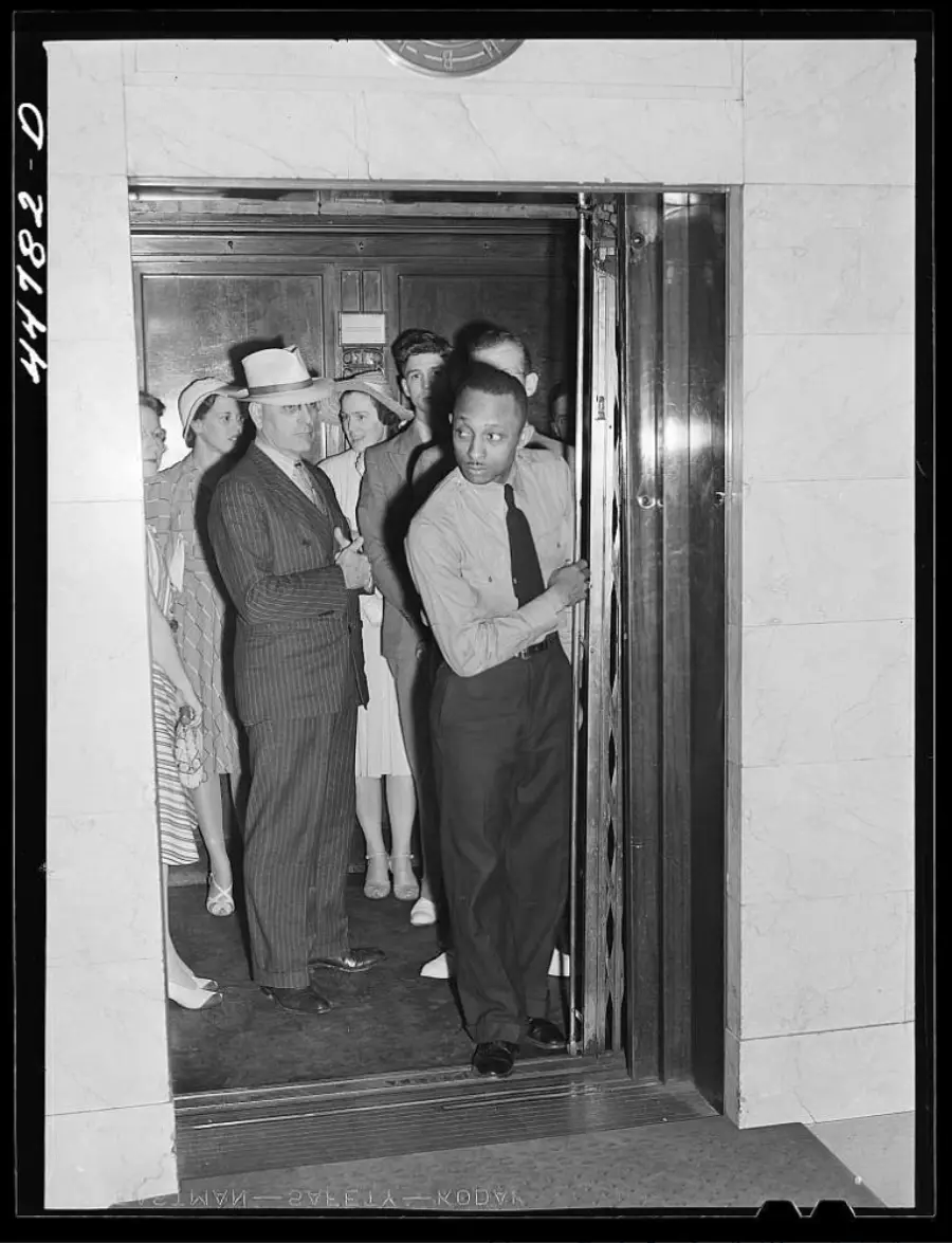 A man operates an elevator while several people, including men in suits and women in dresses and hats, wait to enter. The scene appears to be from the early 20th century.