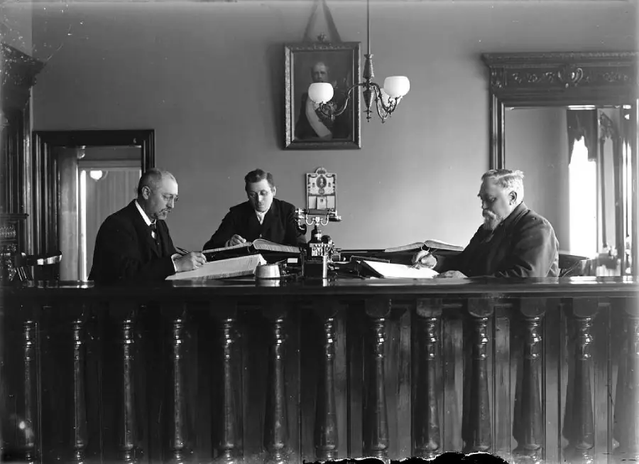 Three men in suits sit and write at a large wooden desk in an ornate office with tall mirrors, a lamp, and a portrait on the wall behind them.